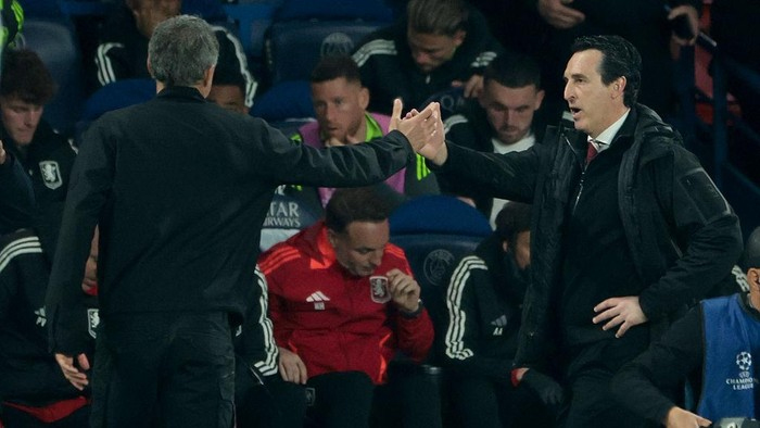 Two football managers on the sideline exchange a handshake during a match.