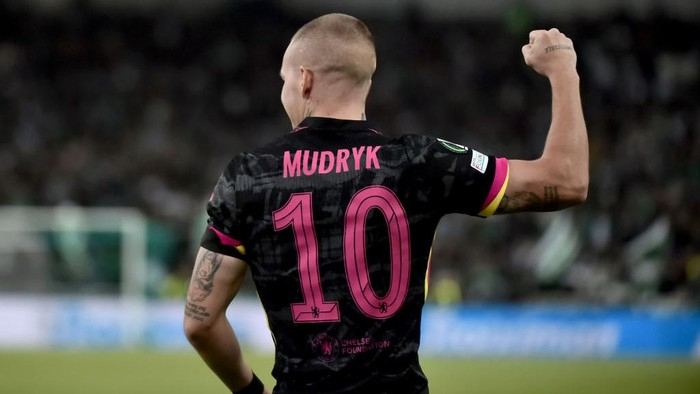 Back view of a soccer player wearing a black Chelsea jersey with pink number 10 and the name Mudryk, fist raised in the air.