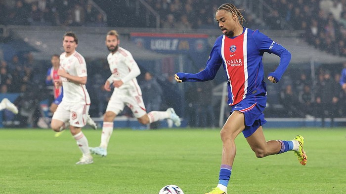Soccer player in a blue-red PSG jersey dribbles the ball on a green field as white-uniform opponents pursue in the background.