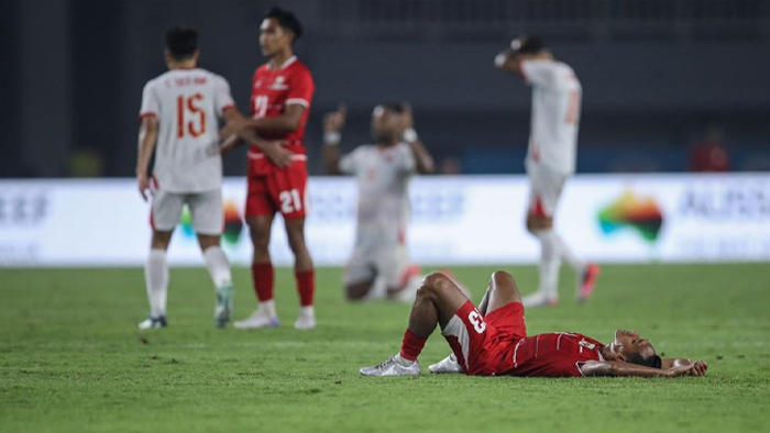 Soccer player in red kit lying on the pitch with arms stretched, while teammates and opponents stand in the background.
