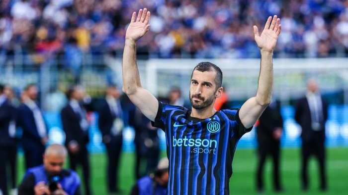 Football player in blue and black striped jersey raises both hands on the field, with blurred crowd behind him.