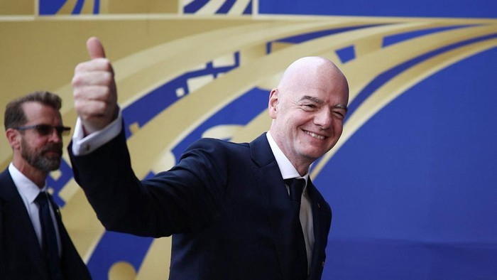 Smiling bald man in a dark suit giving a thumbs-up at a formal event, with a blue and gold backdrop behind him.