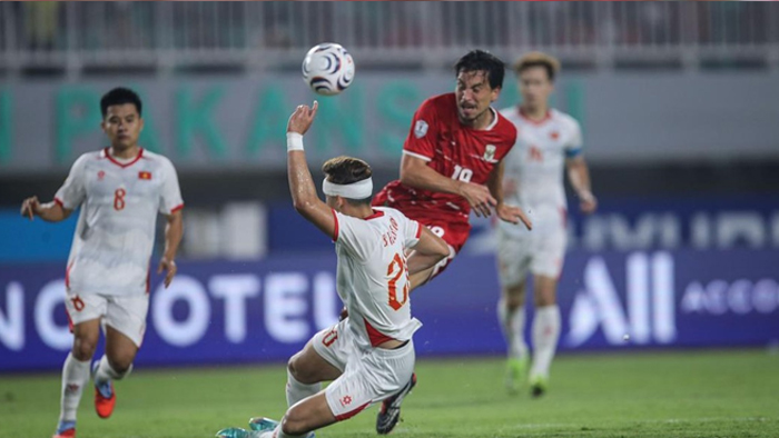 Soccer players in white and red compete for the ball on a grass field; a white-shirted player slides while a red-shirted defender leaps to reach the ball.
