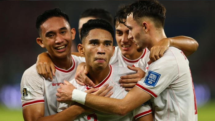 Four soccer teammates in white uniforms celebrate a goal, arms around each other on the field.