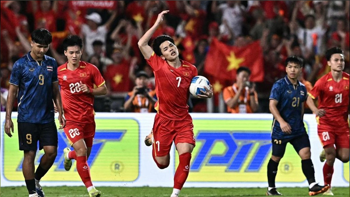 Soccer player in red (number 7) jumps with ball as teammates and opponents run on a pitch with Vietnamese flags in the stands behind them.