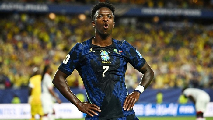 Brazilian footballer in blue Brazil kit #7 celebrates on the field with hands on hips and shouting relief, stadium crowd in the background