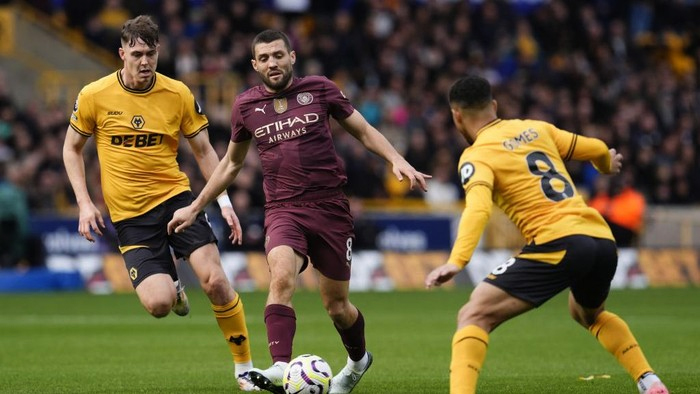 Manchester City player in maroon kit dribbles the ball as two Wolverhampton players in yellow challenge during a match on a green field.