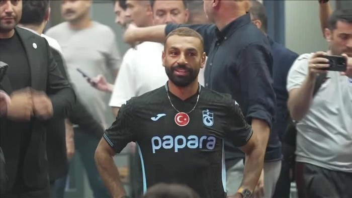 Footballer in a black jersey with 'papara' sponsor and Turkish flag crest posing with hands on hips among fans at a crowded indoor event.