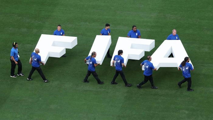 Group of people in blue shirts carry large white 3D letters spelling FIFA across a green field.