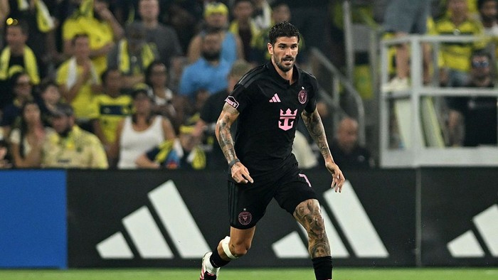 Soccer player in a black kit with pink accents runs on the field during a match, with a crowd in yellow in the stands behind him.