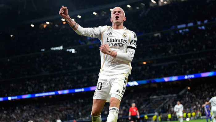 Soccer player in a white Real Madrid kit celebrates with an arm raised on the field, stadium lights in the background.