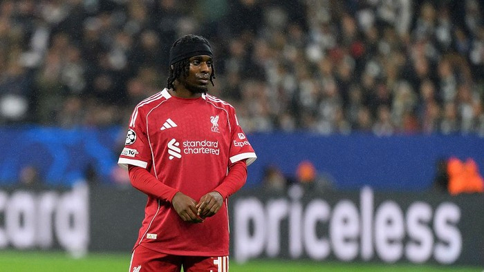 Liverpool FC player in a red home kit standing on the pitch during a match, hands clasped with a blurred crowd behind him.
