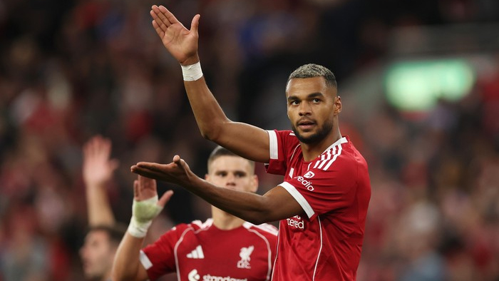 Professional soccer player in a red kit raises his hand to acknowledge fans during a match.