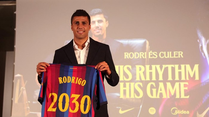 Man in a suit holding up a blue and maroon striped jersey that reads 'RODRIGO 2030' on stage, with a large screen in the background.