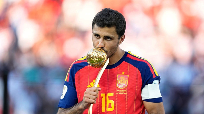 Soccer player in a red Spain jersey (#16) kissing a golden trophy on a ceremonial staff during a celebration.