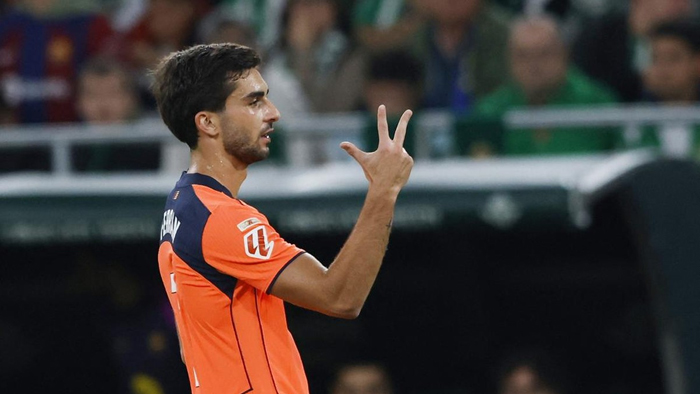 Soccer player in an orange jersey raises three fingers in celebration on the field, with blurred spectators in the background.