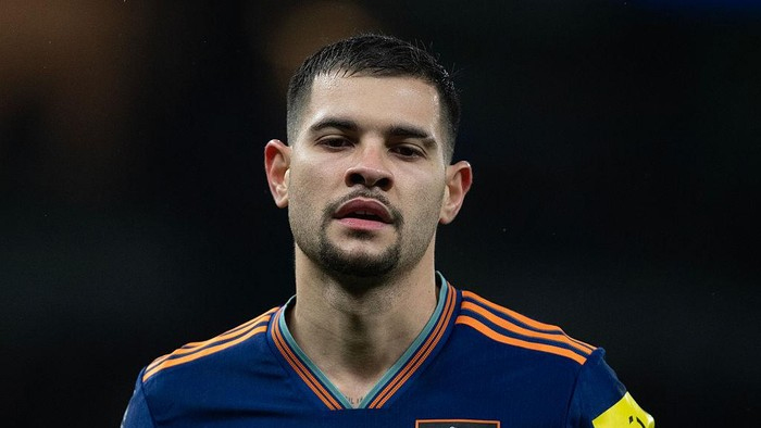 Close-up of a male soccer player in a blue jersey with orange stripes, looking ahead; yellow captain armband visible on his sleeve.