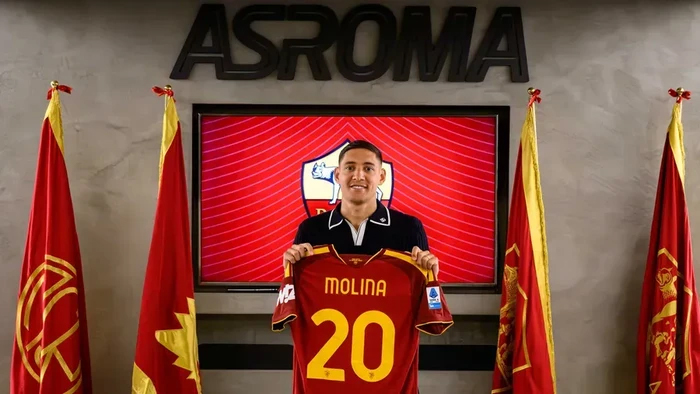 Soccer player Molina holds up a red Roma jersey #20 in front of an AS Roma backdrop with flags on both sides and a red screen behind him.
