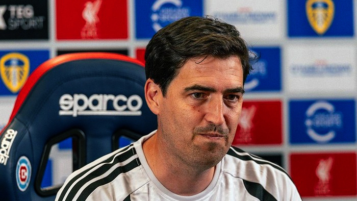 Man in a white sports shirt seated in a blue Sparco chair during a press conference with sponsor banners behind him