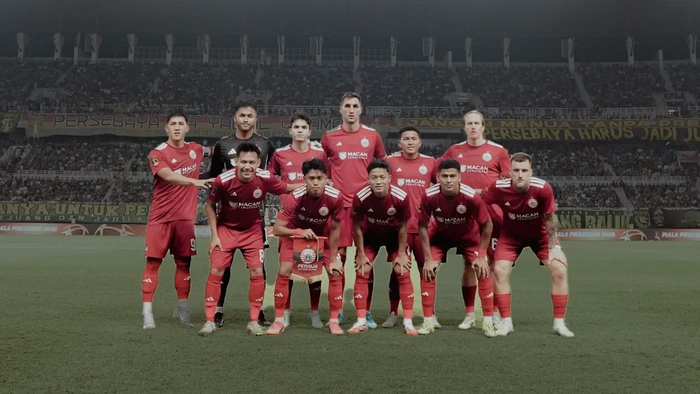 Red-jersey soccer team posing for a group photo on the field in a packed stadium, lineup ready for kickoff.