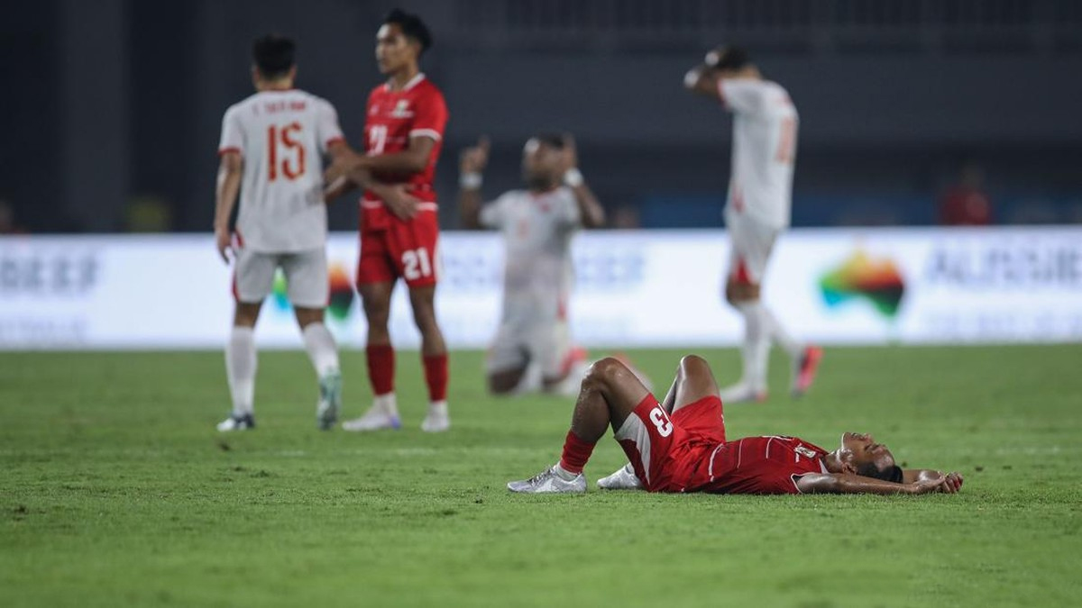 Soccer player in red lying on his back on the grass after apparent injury as teammates stand in the background.