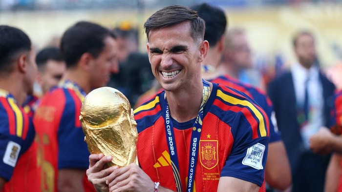 Footballer in a red-and-yellow Spain jersey grins while holding a golden World Cup trophy.