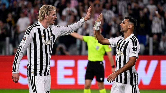Two Juventus players in black-and-white striped kits high-five on the soccer pitch with a cheering crowd behind them.