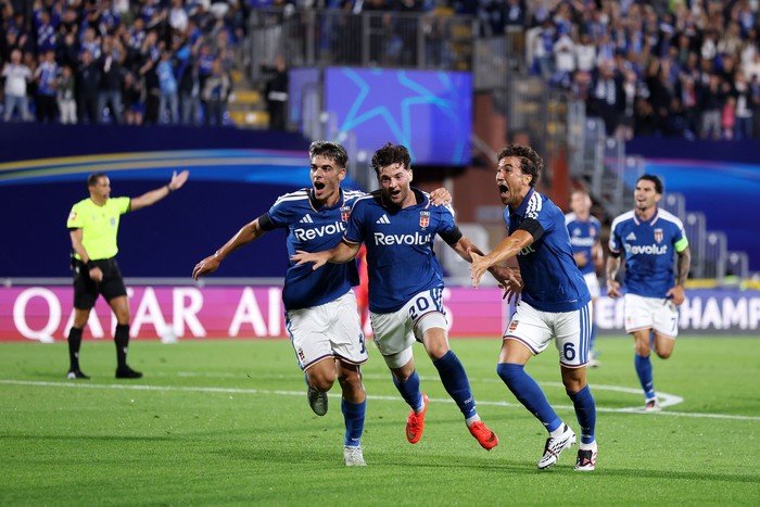 Three Barcelona players celebrate a goal on the pitch, blue home kits, green field, and cheering fans in the stands behind them.