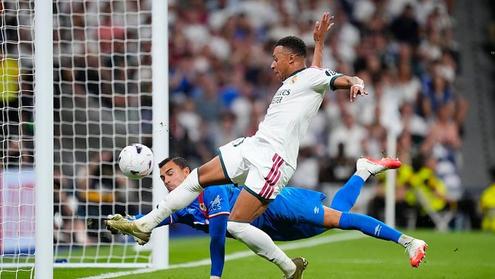 Soccer scene: forward from white team shoots toward the goal while goalkeeper in blue dives to block the ball near the net.