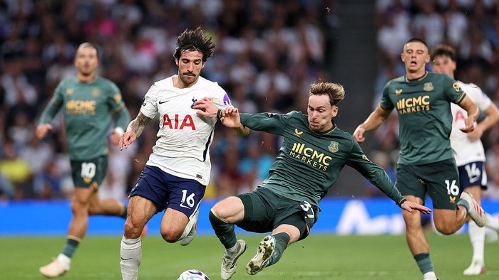 Two soccer players battle for the ball during a match; one in white Spurs kit and one in green kit, with a crowd in the stands behind them.