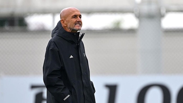 Bald man in a black training jacket standing on a sports field, looking to the right.