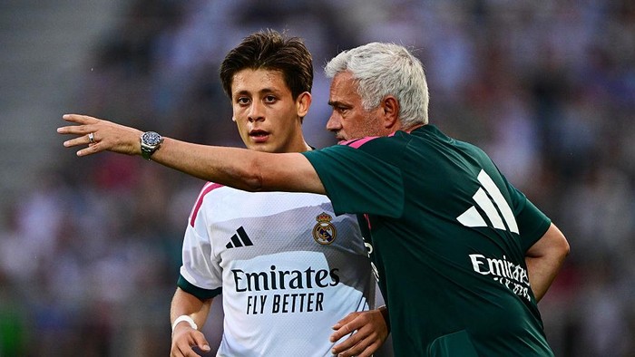 Real Madrid youth player in a white training jersey listens as a coach in green gestures and points during a training session, sponsor logo visible on jersey and coach's back.