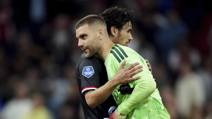 Two soccer players hug on the field, one in a dark jersey and the other in a lime-green goalkeeper kit, with a blurred crowd in the background.