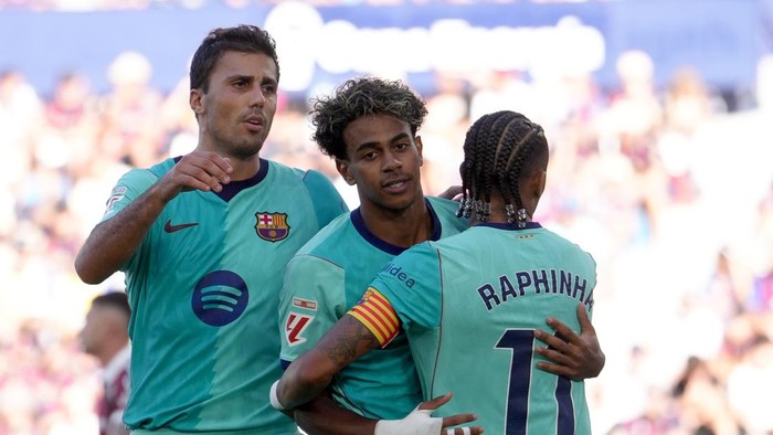 Two Barcelona players hug on the field after a match, one with RAPHINHA 7 on his jersey and captain's armband visible.