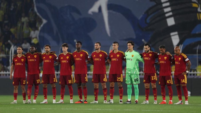 Football team in red jerseys stands in a line on the pitch, goalkeeper in green, before a match, hands linked at the backs of teammates