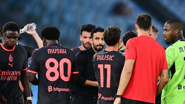 Group of soccer players in black jerseys with pink numbers 80 and 11 stand together on the field after a game; one holds a water bottle/handshake vibe visible.