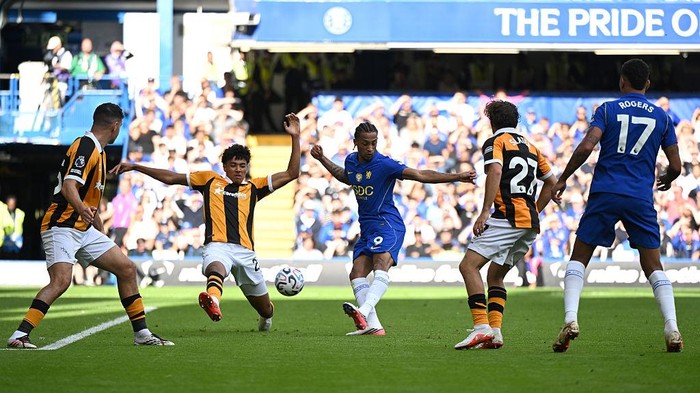 Blue team player in blue kit controls the ball as opponents in yellow-black stripes close in on a sunny pitch to defend.