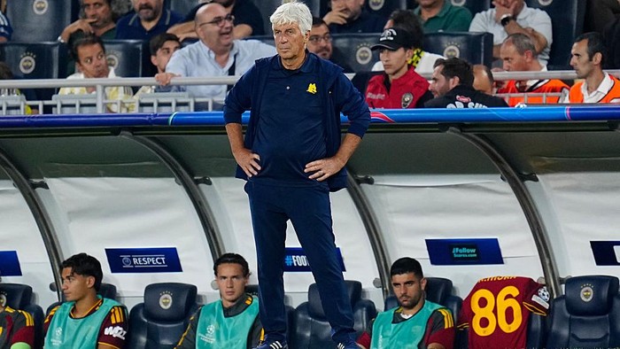 Football coach stands with hands on hips beside players on the bench during a match on the sideline.