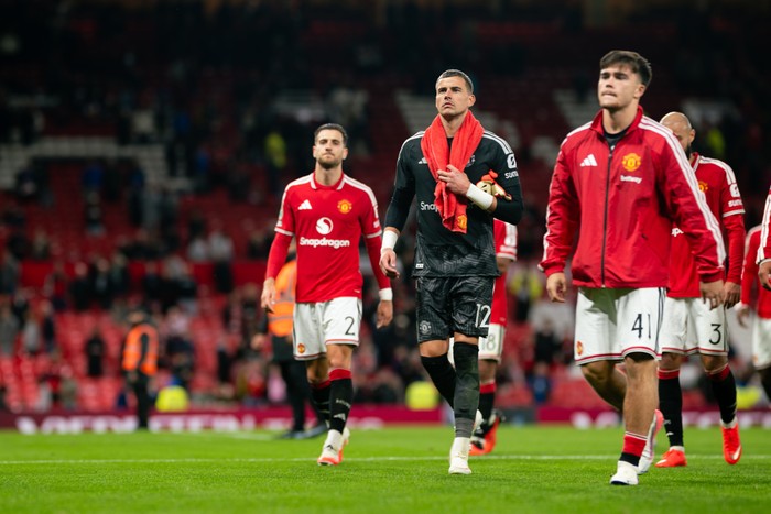 Manchester United players walk off the pitch after a match; goalkeeper in black kit with a red towel over his shoulders in front.