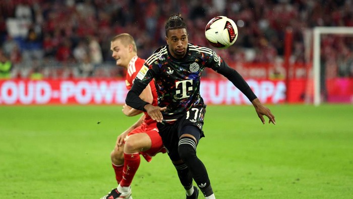 Soccer player in a dark jersey chases a ball midair during a match, with a red-clad opponent nearby on the green field.