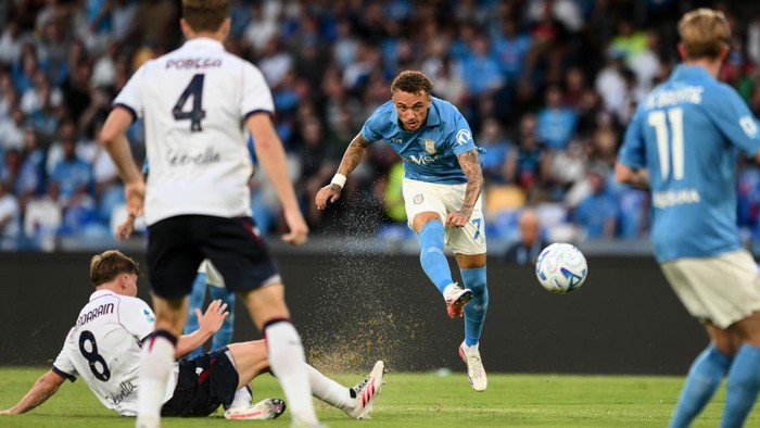 Soccer player in a light blue kit kicks the ball mid-air as opponents in white attempt a challenge on a green pitch with a cheering crowd in the stands.