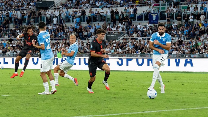 Soccer players compete for the ball on a green pitch as a crowd watches from the stands.