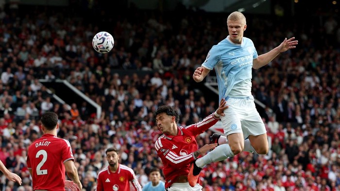 Two football players jump for the ball during a high‑intensity Manchester derby with a packed crowd in the background.