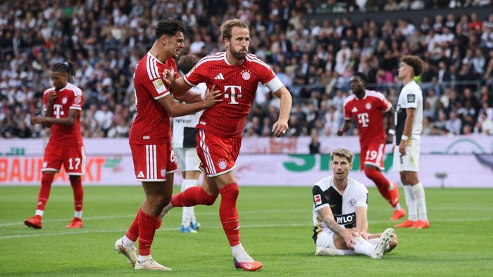 Bayern Munich players in red celebrate on the football pitch while an opposing player sits on the grass.