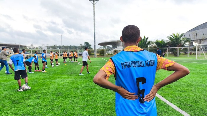Back view of a soccer player in a blue training bib reading 'Papa Football Academy', standing on a green field with teammates practicing in the distance.