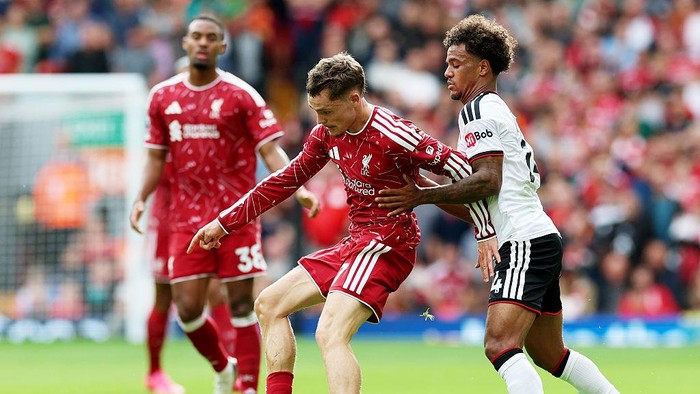 Liverpool player in a red jersey challenges for the ball against a defender in white and black kit during a match on a green field.