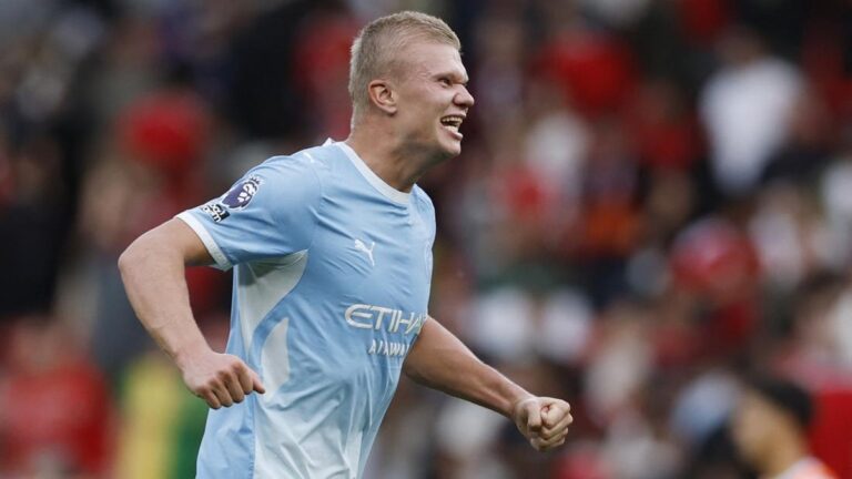 Soccer player in a light blue Manchester City jersey cheering with clenched fists on the pitch during a match, crowd in the background.