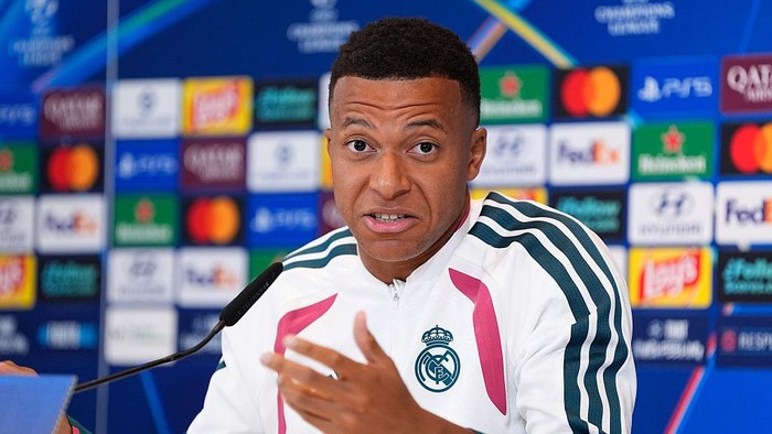 Footballer wearing a Real Madrid jacket speaks at a press conference with sponsor logos in the backdrop.