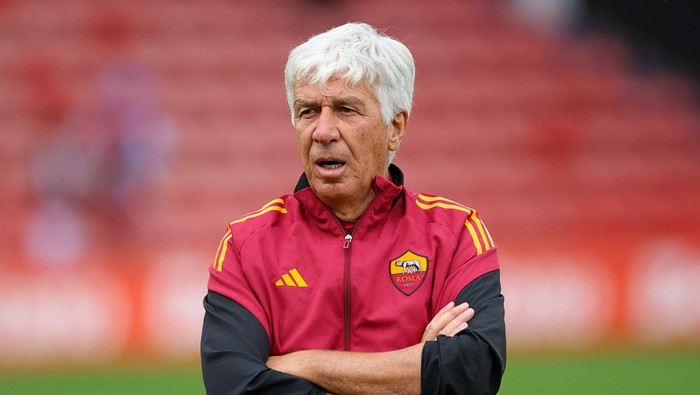 Football coach wearing a red Roma training jacket standing with arms crossed on a stadium field.