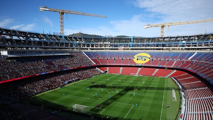 A large soccer stadium under construction with cranes overhead, crowded stands, and players on a green pitch.